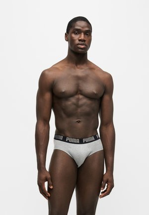 Homme musclé debout devant un fond uni, portant un slip gris Puma avec une ceinture noire.