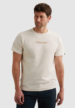 Mężczyzna z krótkimi brązowymi włosami, ubrany w beżowy T-shirt z krótkim rękawem z napisem "PME LEGEND" oraz ciemnoniebieskie spodnie, stojący na jednolitym tle.