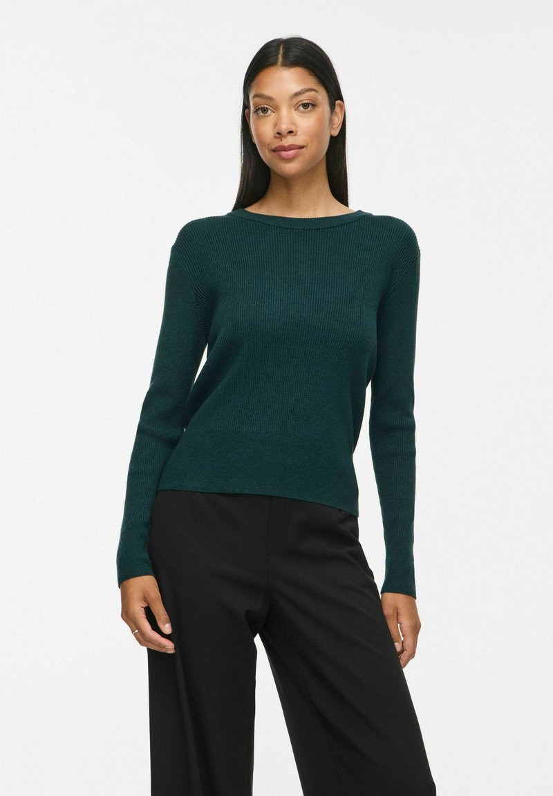 Femme aux longs cheveux foncés portant un pull côtelé vert foncé et un pantalon noir, debout devant un fond blanc uni.