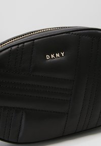 Pochette DKNY en cuir noir avec couture matelassée et fermeture éclair dorée sur le dessus, affichant le logo de la marque en lettres dorées.