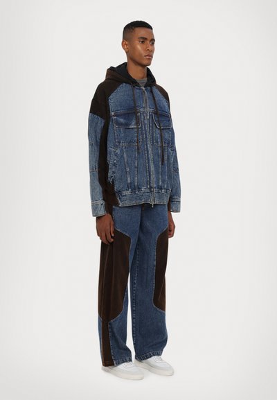 Denim jacket with brown accents, two chest pockets, zip closure. Paired with wide-leg jeans featuring brown panels and white sneakers.