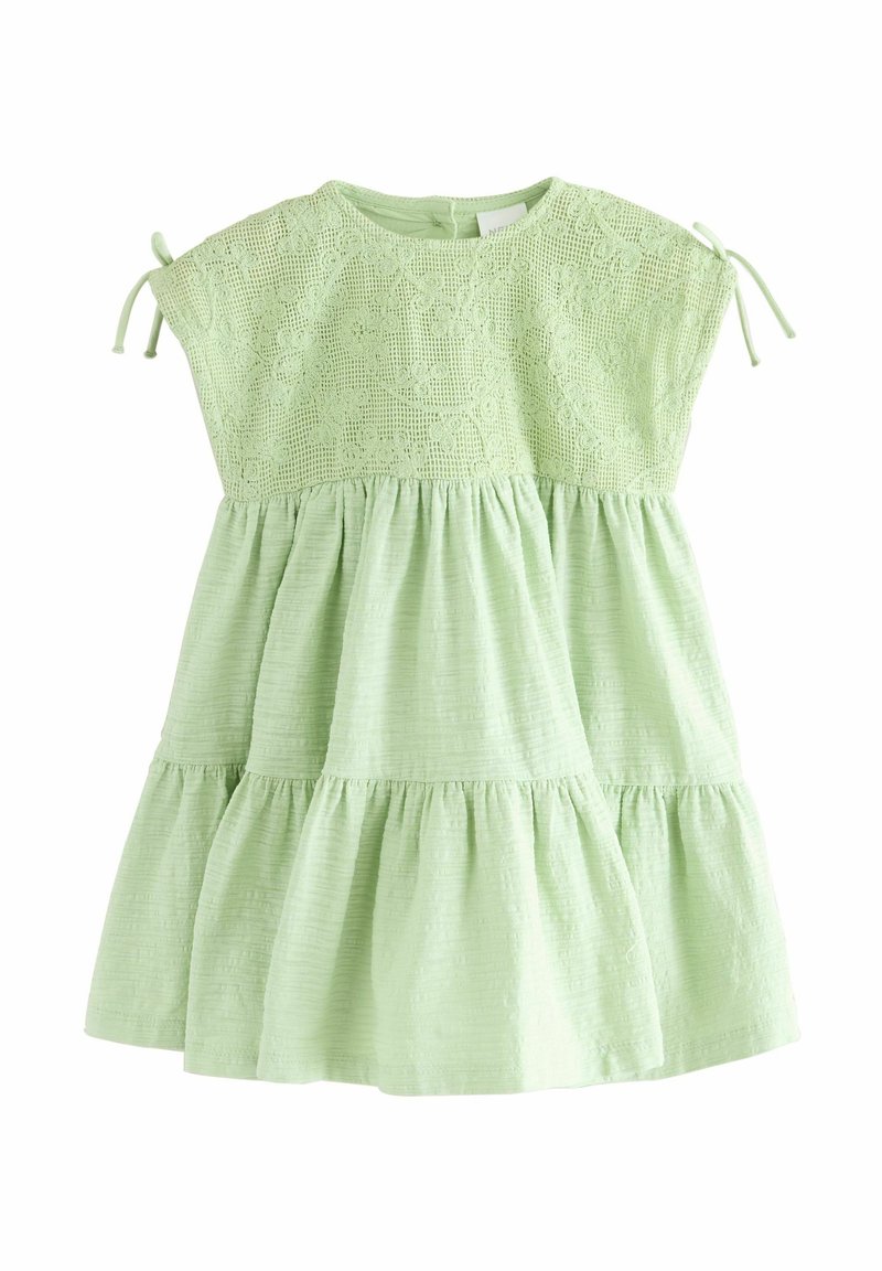 Lichtgroene jurk met een textuur kant top, korte mouwen en geplooide lagen bij de taille. Heeft strikaccenten bij de schouders.