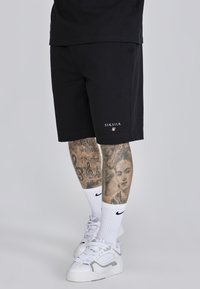 Čierne bavlnené šortky s logom na ľavom stehne, v kombinácii s bielymi ponožkami Nike a bielymi teniskami, zdôrazňujúce detailné tetovania na nohách.