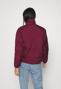 Burgundy puffjacka med bladmönster, hög krage, elastiska ärmar och en texturerad finish. Bärs med ljusblå, högmidjade jeans.