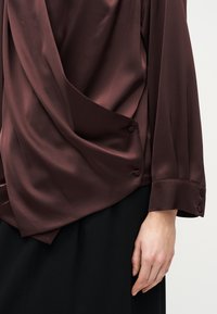 Σκούρα καφέ σατέν μπλούζα με κουμπιά στο μανίκι και στο πλάι, φορεμένη με μαύρη φούστα πάνω σε ανοιχτό φόντο.