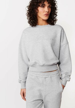 Kobieta o kręconych włosach, ubrana w jasnoszarą bluzę z okrągłym dekoltem i pasujące spodnie dresowe, stojąca z jedną ręką w kieszeni.