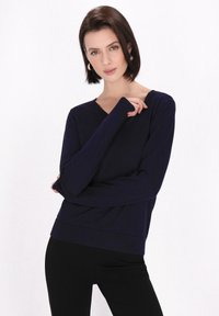 Maglione a maniche lunghe blu navy con scollo a v, realizzato in tessuto morbido, con polsini e orlo a costine, indossato sopra pantaloni neri, con dettagli di stile minimi.