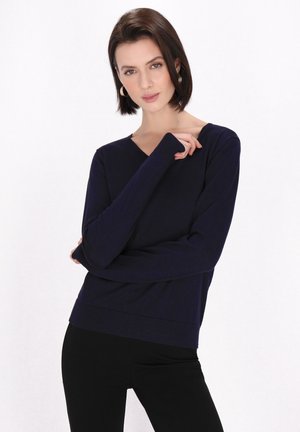 Mørkeblå langærmet v-udskåret sweater lavet af blødt stof med ribbede manchetter og kant, båret over sorte bukser, med minimalt stylingdetaljer.