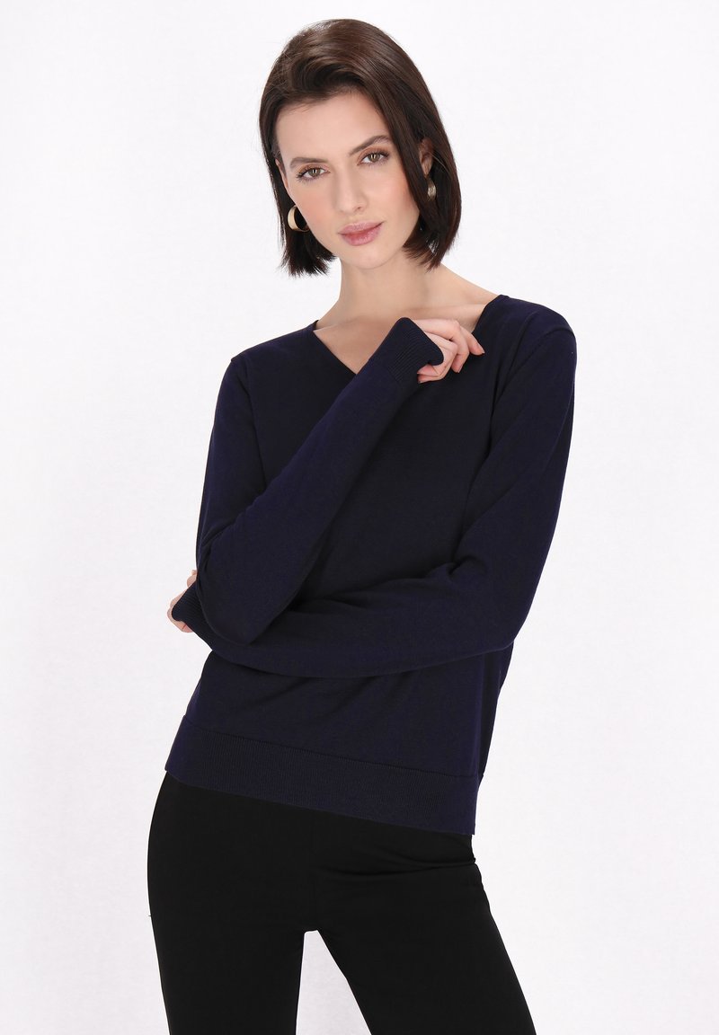 Maglione a maniche lunghe blu navy con scollo a v, realizzato in tessuto morbido, con polsini e orlo a costine, indossato sopra pantaloni neri, con dettagli di stile minimi.