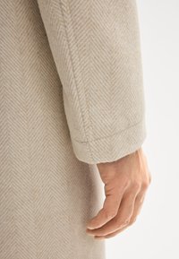 Beiger Mantelärmel mit Fischgrätmuster aus einer strukturierten Stoff; zeichnet sich durch saubere Nähte und einen umgeschlagenen Manschettenrand aus. Menschliche Hand sichtbar.