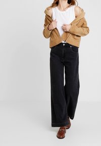 Brązowa kurtka z kordurowego materiału, krótka, na białym topie na ramiączkach, w połączeniu z szerokimi czarnymi dżinsami i brązowymi butami na slip-on. Prosty, codzienny strój.