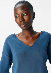 Donna con pelle scura che indossa un maglione blu a scollo a V, che guarda a sinistra su uno sfondo bianco semplice.
