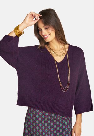 Lächelnde Frau mit braunen Haaren trägt einen dunkelvioletten Pullover, mehrere goldene Halsketten, goldene Armreifen und einen Rock mit geometrischem Muster.