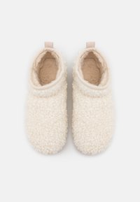 Pantuflas de felpa de color crema con un exterior suave y texturizado, diseño minimalista y una pequeña lengüeta a juego en el talón.