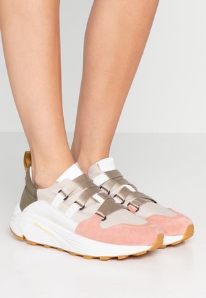 Nogi w beżowych, różowych i oliwkowozielonych sneakersach z grubymi białymi podeszwami i wieloma szerokimi paskami, stojące na jasnoszarej powierzchni.