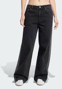 Schwarze weit geschnittene Jeans aus Denim, mit hoher Taille, Fronttaschen und seitlichen Streifen. Getragen mit weißen Sneakers.