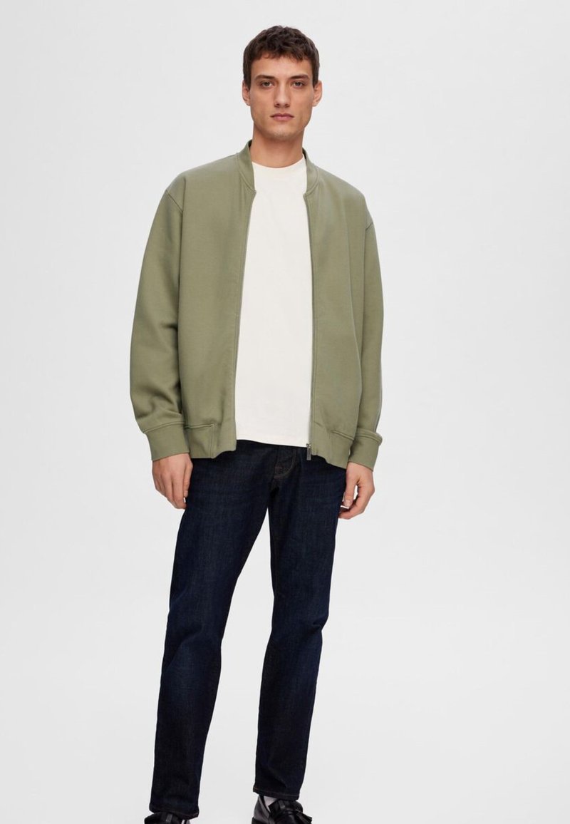 Veste bomber zippée vert olive avec des poignets et un ourlet côtelés, portée sur un t-shirt blanc, associée à un jean bleu foncé et des chaussures noires.