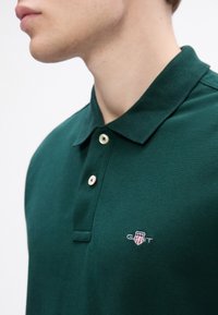 Mees, kes kannab tumeroheline polo-särki, millel on kaks valget nuppu ja väike tikand GANT logo rinnal, valge tausta ees.