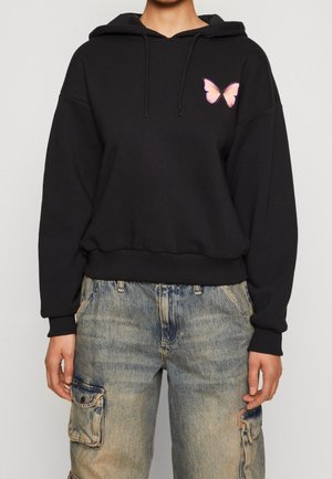 Person trägt einen schwarzen Hoodie mit einem kleinen rosa Schmetterling auf der Brust und ausgewaschene blaue Cargo-Jeans.