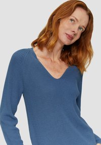 Blauer Strickpullover mit V-Ausschnitt, langen Ärmeln, strukturiertem Stoff und gerippten Details entlang des Ausschnitts und der Seiten.