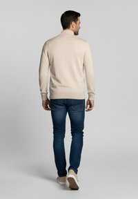 Suéter beige claro con cuello alto, diseño ajustado, combinado con jeans azul oscuro y zapatillas claras, visto desde atrás.