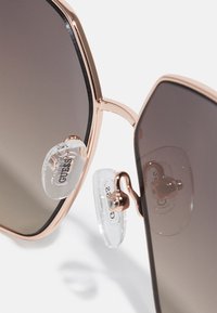 Ochelari de soare roz aurie cu lentile maro cu gradient, având suporturi nazale din silicon transparent și un design al ramei geometric.