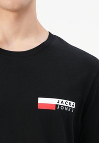 Zwarte katoenen t-shirt met een logo op de linkerkant van de borst, witte tekst en een rood-witte rechthoekige accent. Ronde hals, korte mouwen.