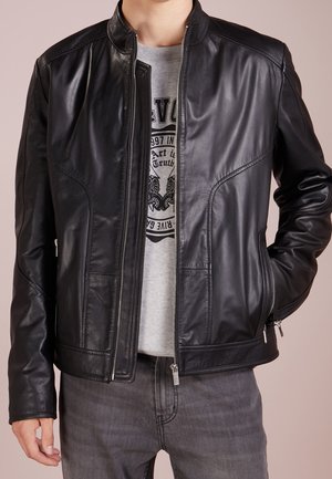 Schwarze Lederjacke mit Stehkragen, Reißverschluss vorne und seitlichen Taschen. Sie hat eine glatte Textur und eine taillierte Passform. Wird über einem grauen T-Shirt getragen.