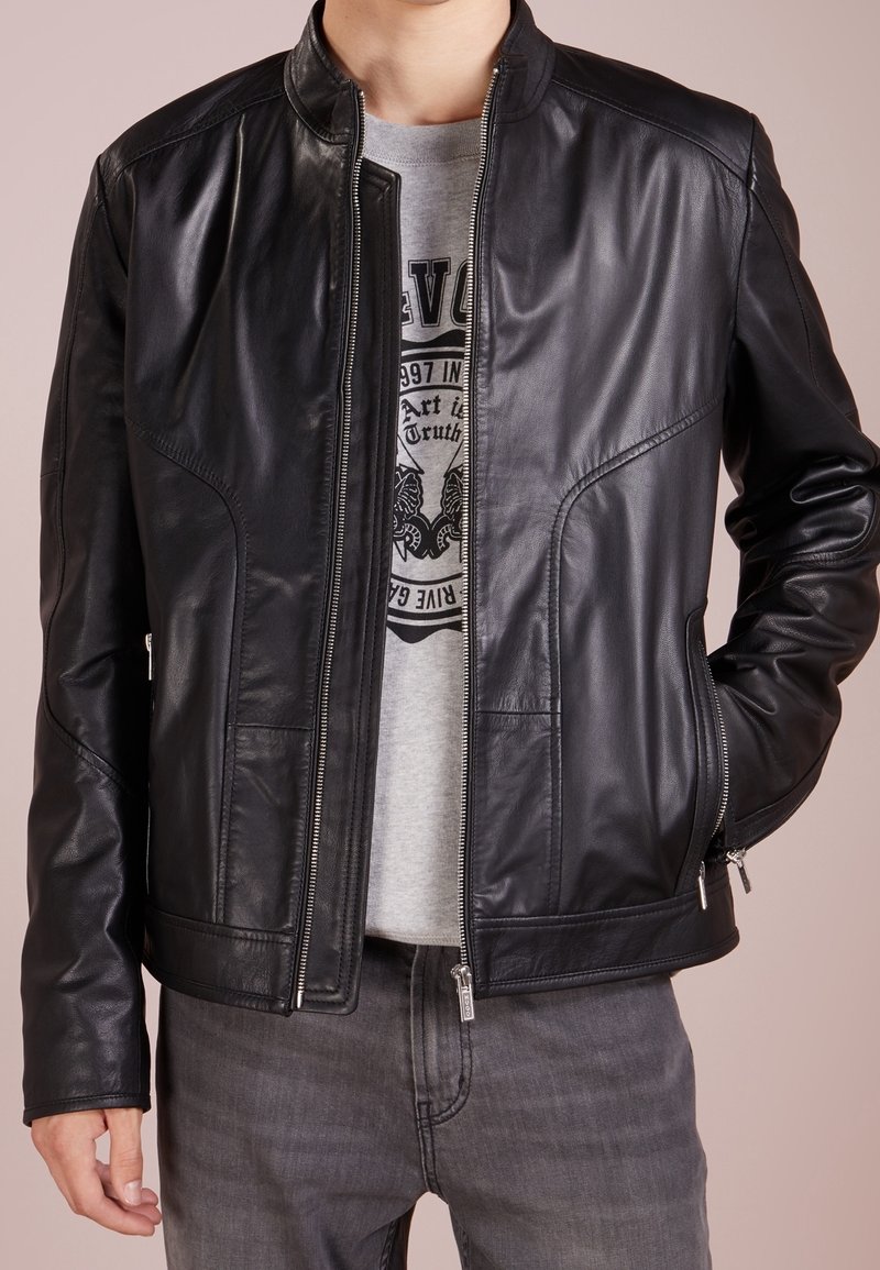 Schwarze Lederjacke mit Stehkragen, Reißverschluss vorne und seitlichen Taschen. Sie hat eine glatte Textur und eine taillierte Passform. Wird über einem grauen T-Shirt getragen.