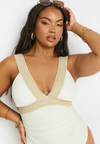 Valkoinen uimapuku, jossa on syvä V-aukko ja rakenteelliset kultaiset yksityiskohdat kauluksessa ja vyötäröllä. Malli poseeraa pitkät tummat hiukset ja korvakkaat korvakorut.