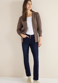 Brązowy prążkowany kardigan na białej koszulce, w zestawieniu z ciemnoniebieskimi jeansami zapinanymi na dwa guziki. Noszone z jasnymi sneakersami, na neutralnym tle.