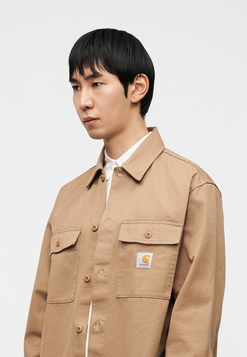 Camicia marrone chiaro con bottoni, dotata di due tasche sul petto, una toppa con il logo sulla tasca sinistra e una vestibilità comoda con colletto classico.