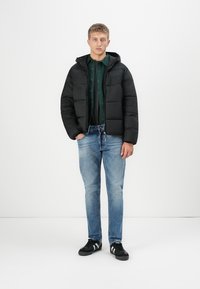 Ung mand står med hænderne i lommerne, iført sort pufferjakke, mørkegrøn skjorte, lyseblå jeans og sorte sneakers med hvide striber.