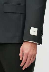 Jachetă gri cu un croi slim, având un etichetă crem cu "Calvin Klein" pe manșetă și o țesătură texturată. Accente cu nasturi pe mânecă.