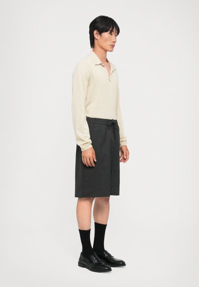 Hellbeige Langarm-Polohemd, graue Shorts mit Kordelzug, schwarze Slipper und knielange schwarze Socken an einem männlichen Modell, Seitenansicht.