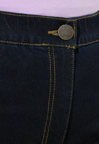 Tmavě modré džíny z denimové látky mají kovové zapínání, žluté švy a hladkou texturu s mírně vybledlým povrchem.