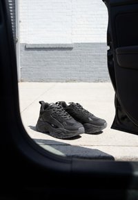 Par sorte sneakers placeret på beton udenfor en bil med en åben dør foran en hvid og grå murstensvæg.