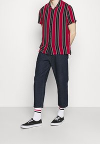 Mężczyzna stojący z rękami za plecami, ubrany w czerwoną i czarną koszulę w pionowe paski na krótkich rękawach, skrócone ciemne dżinsy, białe skarpetki i czarne trampki.