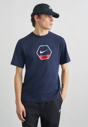 Junger Mann trägt ein marineblaues Nike-T-Shirt mit sechseckigem Logo, schwarze Nike-Kappe und schwarze Hose, schaut nach rechts vor einem schlichten Hintergrund.