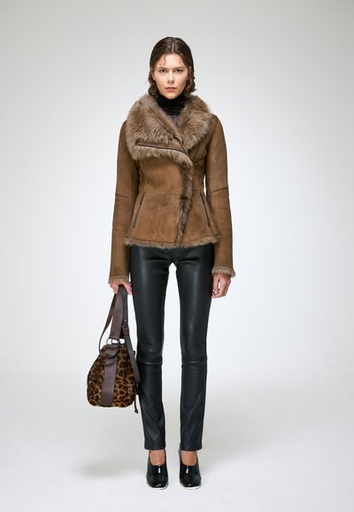 Femme portant une veste marron à col en fourrure, pantalon en cuir noir, chaussures noires, tenant un sac à main à imprimé léopard, debout devant un fond uni.