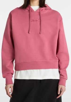 Persoon die een cropped roze hoodie draagt met "vans" geborduurd op de borst, gelaagd over een wit overhemd en gecombineerd met een zwarte broek.