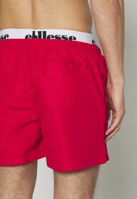 Rote Badehose aus leichtem Material mit einem elastischen Bund, der ein schwarzes Logo trägt. Enthält eine Gesäßtasche mit Klappe.