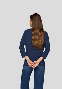 Dunkelblause Baumwollbluse mit dreiviertel Ärmeln, geradem Schnitt und Rundhalsausschnitt, kombiniert mit hellblauen Jeans. Die Haare fallen über die Schultern.