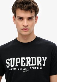 Sort bomulds t-shirt med fed hvid tekst "SUPERDRY" med "PREMIUM SPORTING" under. Crew neck og kortærmet design.