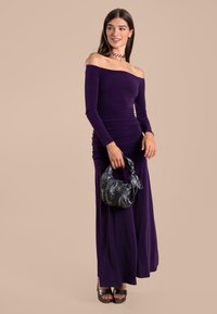 Robe violette décolletée avec des manches longues, des détails froncés et une jupe fluide. Associée à un sac à main noir texturé et des escarpins compensés.