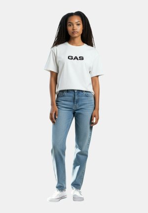 Donna in piedi che indossa una t-shirt bianca con la scritta "GAS", jeans azzurri a vita alta e sneakers bianche su sfondo semplice.