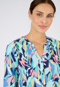 Blusa colorida con un estampado botánico en azul, rosa y verde. Tiene un escote redondo y botones en la parte delantera, con una tela texturizada.