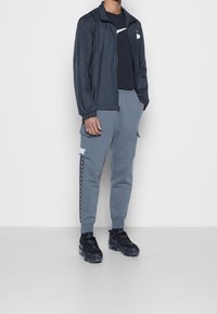 Marine windbreaker jack, zwart T-shirt, grijze sweatpants met zijzakken en wit logo, zwarte sport sneakers, casual fit.