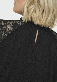 Czarna koronkowa bluzka z wysokim dekoltem, przezroczystymi koronkowymi rękawami i małym zapięciem na guzika z tyłu, prezentująca misternie wykonane wzory kwiatowe.