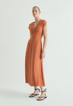 Mujer de pie vistiendo un vestido midi naranja de manga corta con fruncidos y sandalias planas negras con lazada en el tobillo, sobre un fondo claro liso.